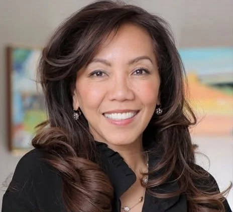 A woman with long brown hair smiling at the camera, wearing a black top and earrings, with a colorful, blurred background.