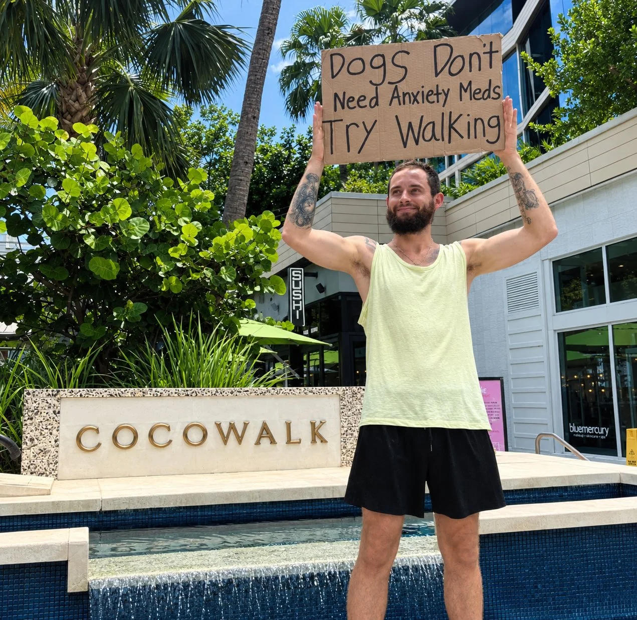 Bearded man holding a sign reading “Dogs Don’t Need Anxiety Meds, Try Walking” outside Cocowalk in Miami.