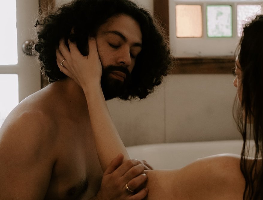 A person with long, dark hair is touching the face of a bearded man with curly hair, both in a close and intimate setting, possibly a bathroom.
