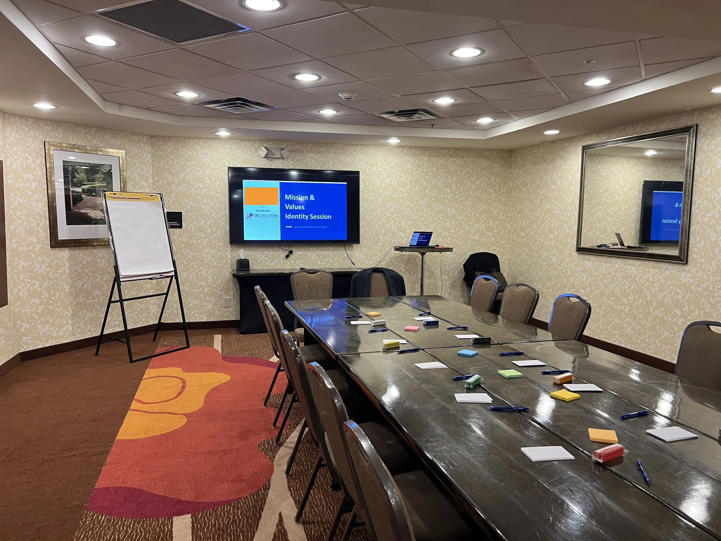 A long wooden table with notecards, post-it notes, and pens on the table. In the front of the room is a white easel and television.