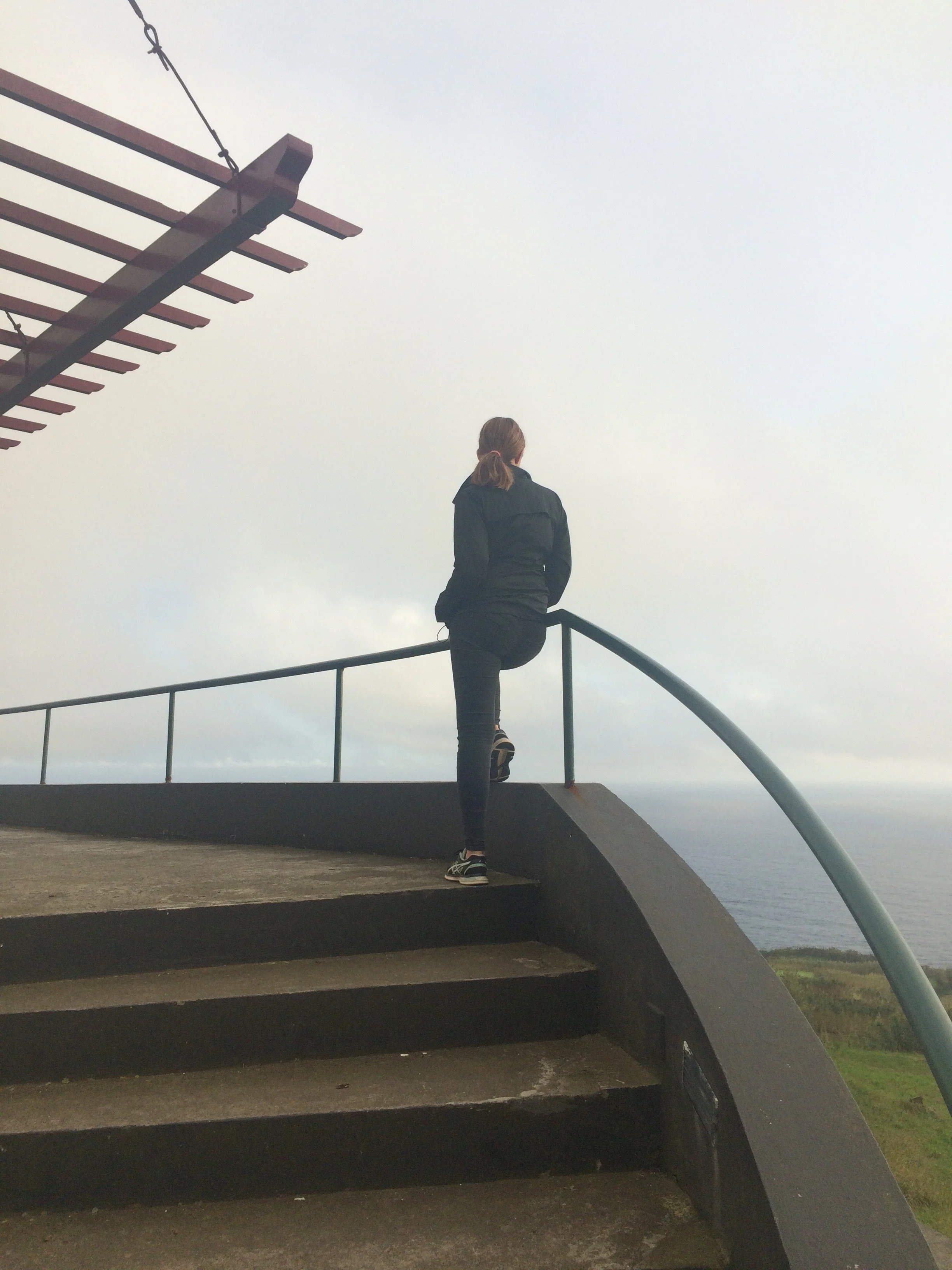 Woman in jeans and a black jacket looking out into nature from the top of a staircase