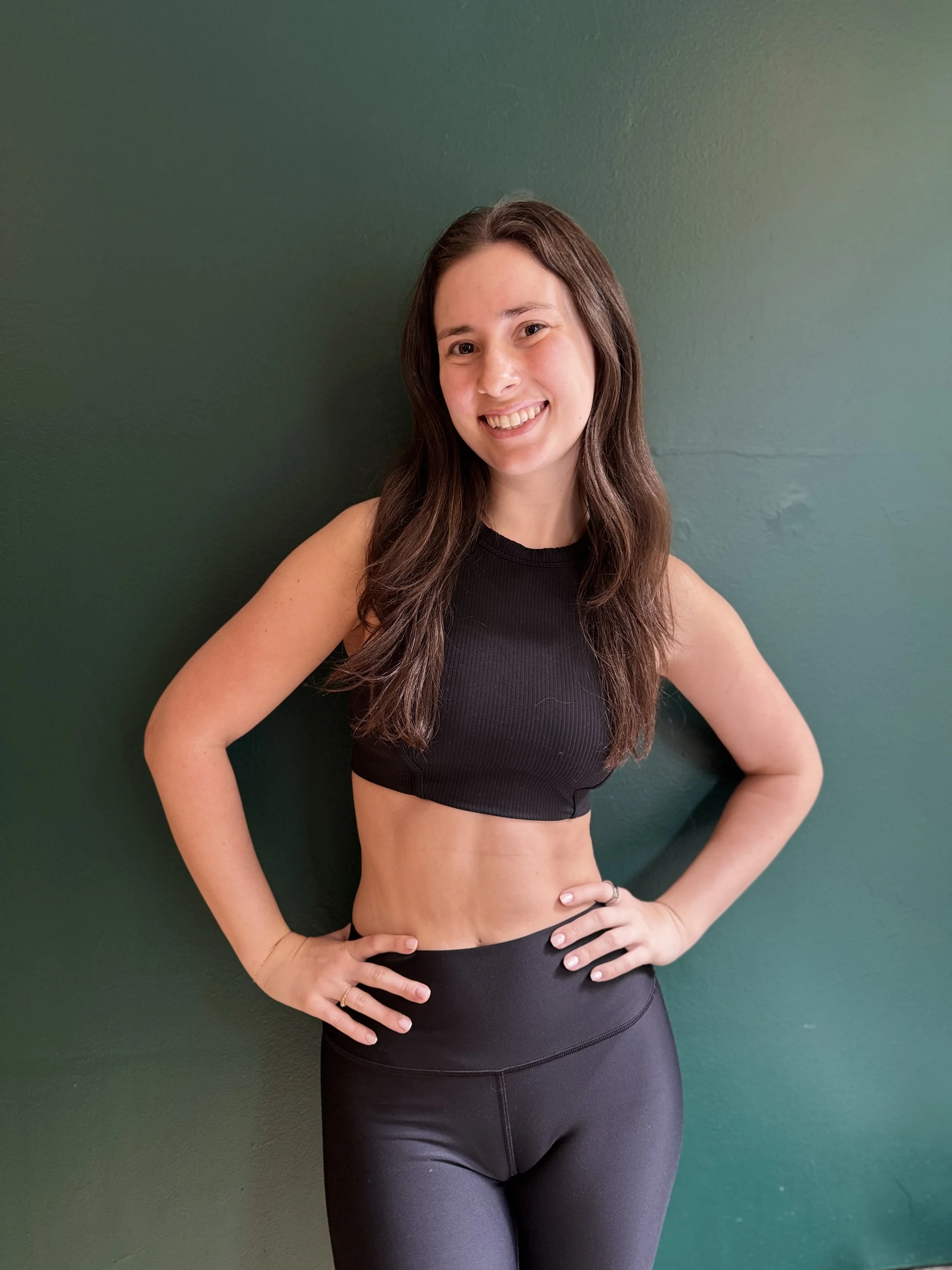 A young woman with long brown hair, smiling, wearing a black sleeveless crop top and black athletic leggings, standing against a green wall.
