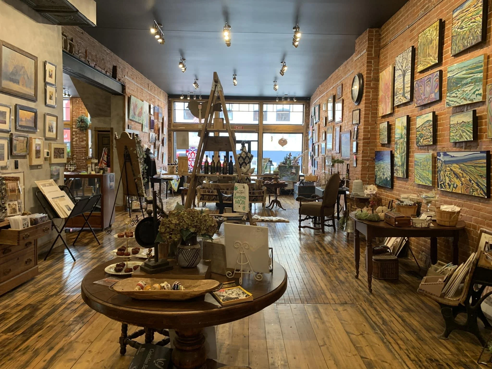 Interior of an art gallery with paintings on brick walls, wooden floors, and display tables, featuring artwork, ceramics, and decorative items.
