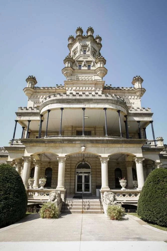 A historic, ornate castle-like building with domed turrets, columns, and balconies, set against a clear blue sky.