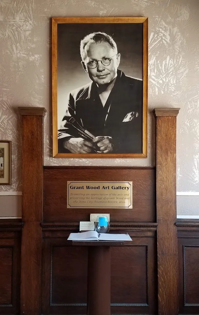 A large black and white portrait of an elderly woman with glasses and short hair, holding a paintbrush, hanging on a wall inside a gallery. Below the portrait, there is a sign that reads 'Grant Wood Art Gallery,' and a small table with an open book and a glass of water.