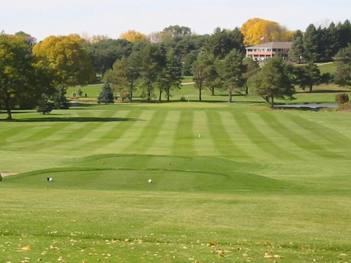 A golf course with a neatly mowed green fairway, surrounded by trees with autumn foliage, and a large house in the background.