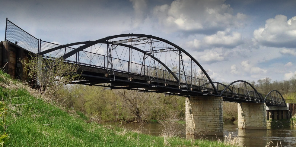 Old metal bridge with arches spanning over a river, surrounded by greenery and under a cloudy sky.