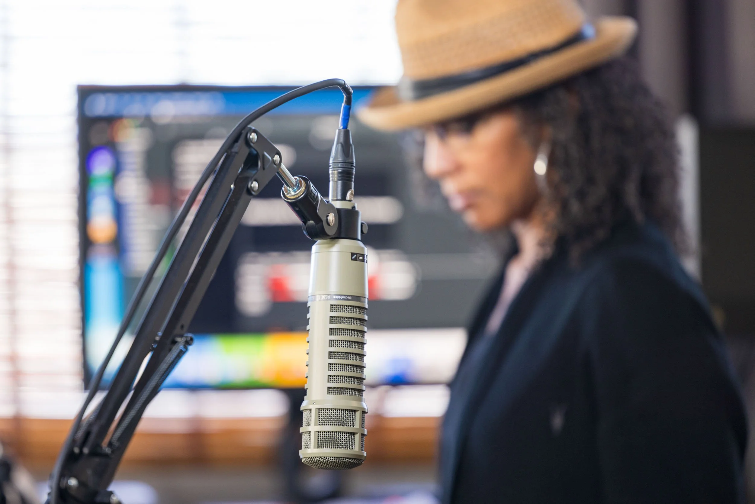 Close-up of a professional condenser microphone hanging in front of a woman with curly hair wearing a hat, in a recording studio.