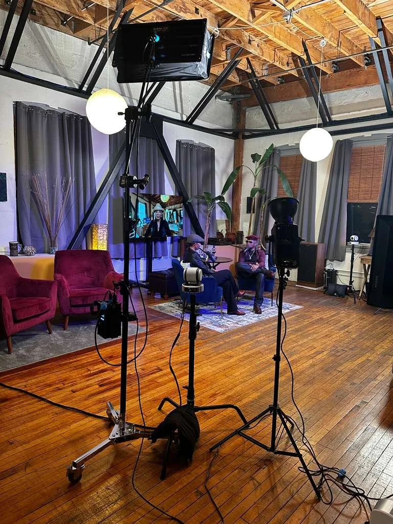 Indoor TV studio or podcast set with two people sitting on chairs, microphones, lighting equipment, a large flat-screen TV, couches, and decorative plants.