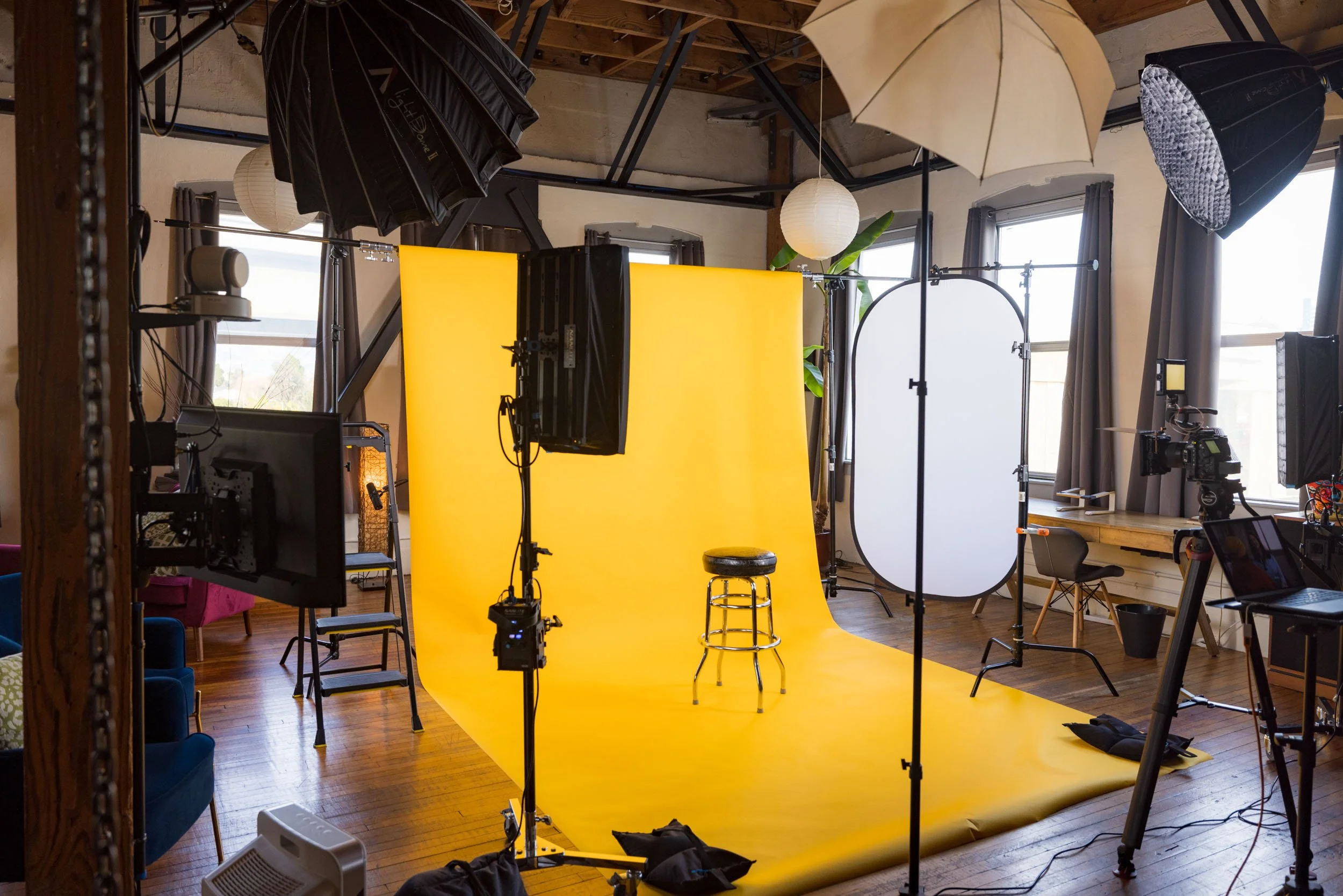 A photography studio with a yellow backdrop, various studio lights, umbrellas, and cameras set up for a photo shoot in a room with wooden floors and large windows.