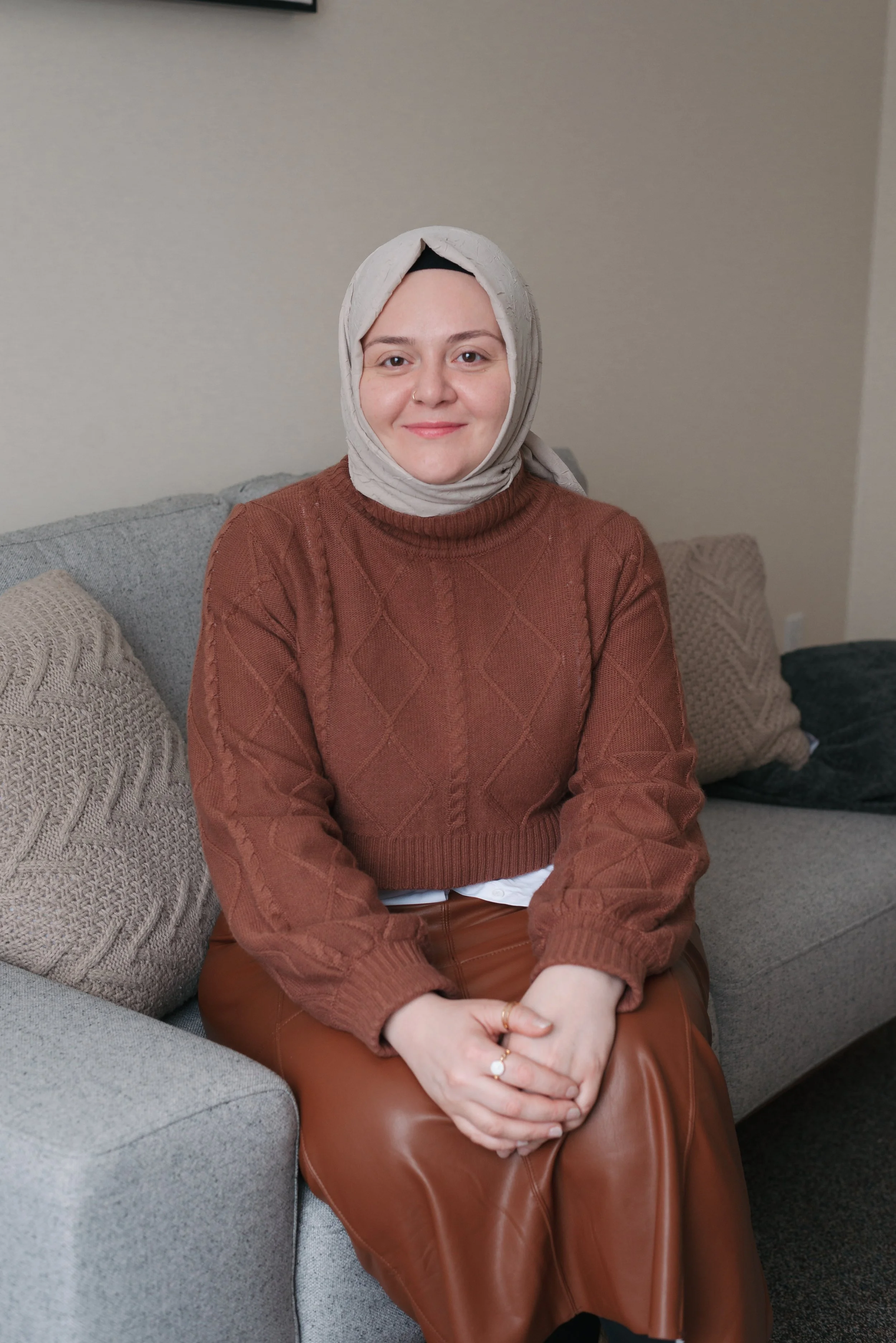 A woman wearing a gray hijab and a teal dress sitting on a gray couch with a decorative pillow behind her. She has a light-colored long-sleeve shirt underneath and is accessorized with multiple necklaces, bracelets, and rings. Behind her, there are string lights and a large leafy plant.