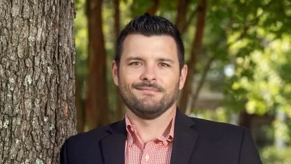 A portrait of a man with short dark hair, a beard, wearing a suit jacket, dress shirt, and bowtie, standing outdoors with trees in the background.