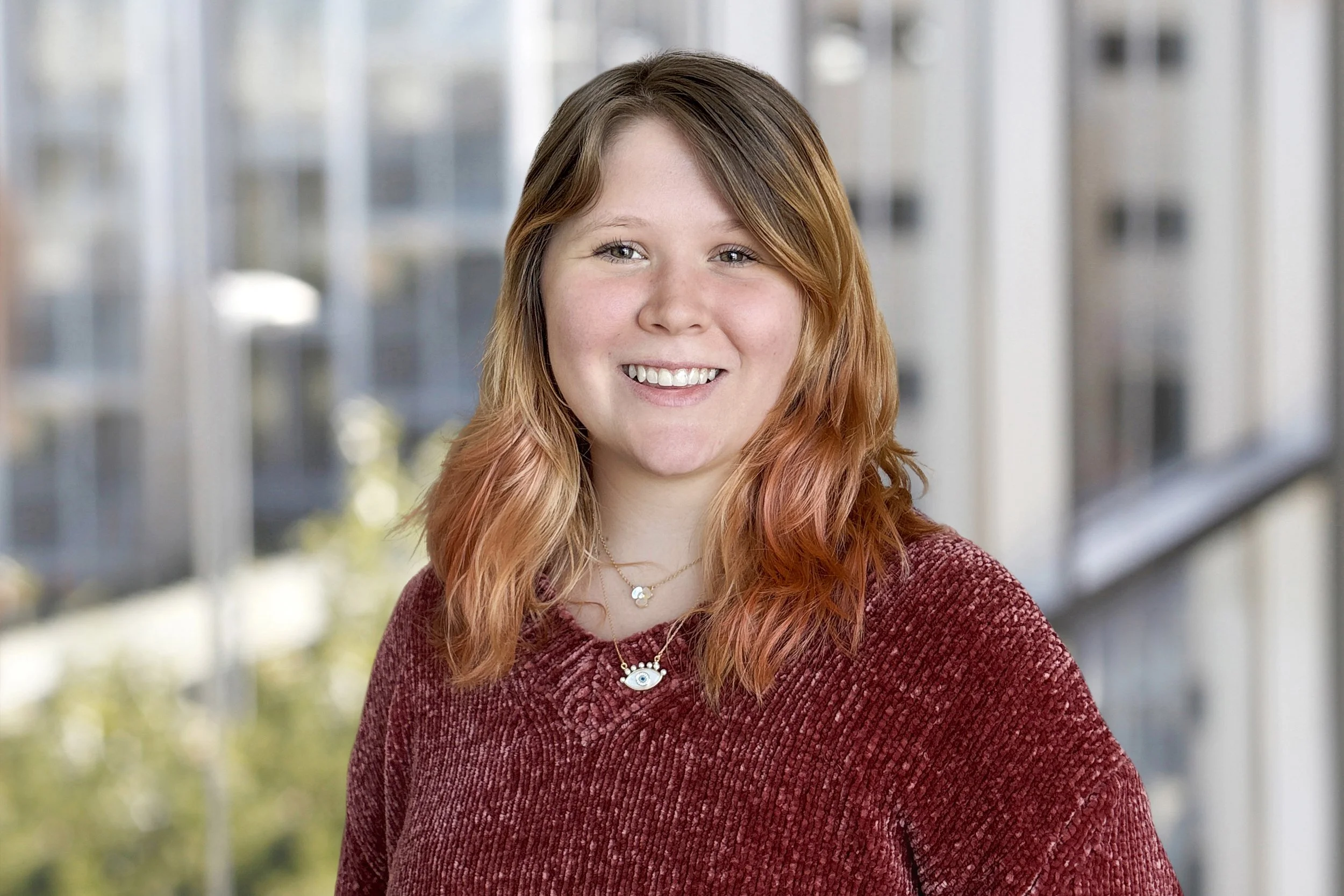 A woman outdoors with trees in the background, smiling and wearing a sweater and necklace.