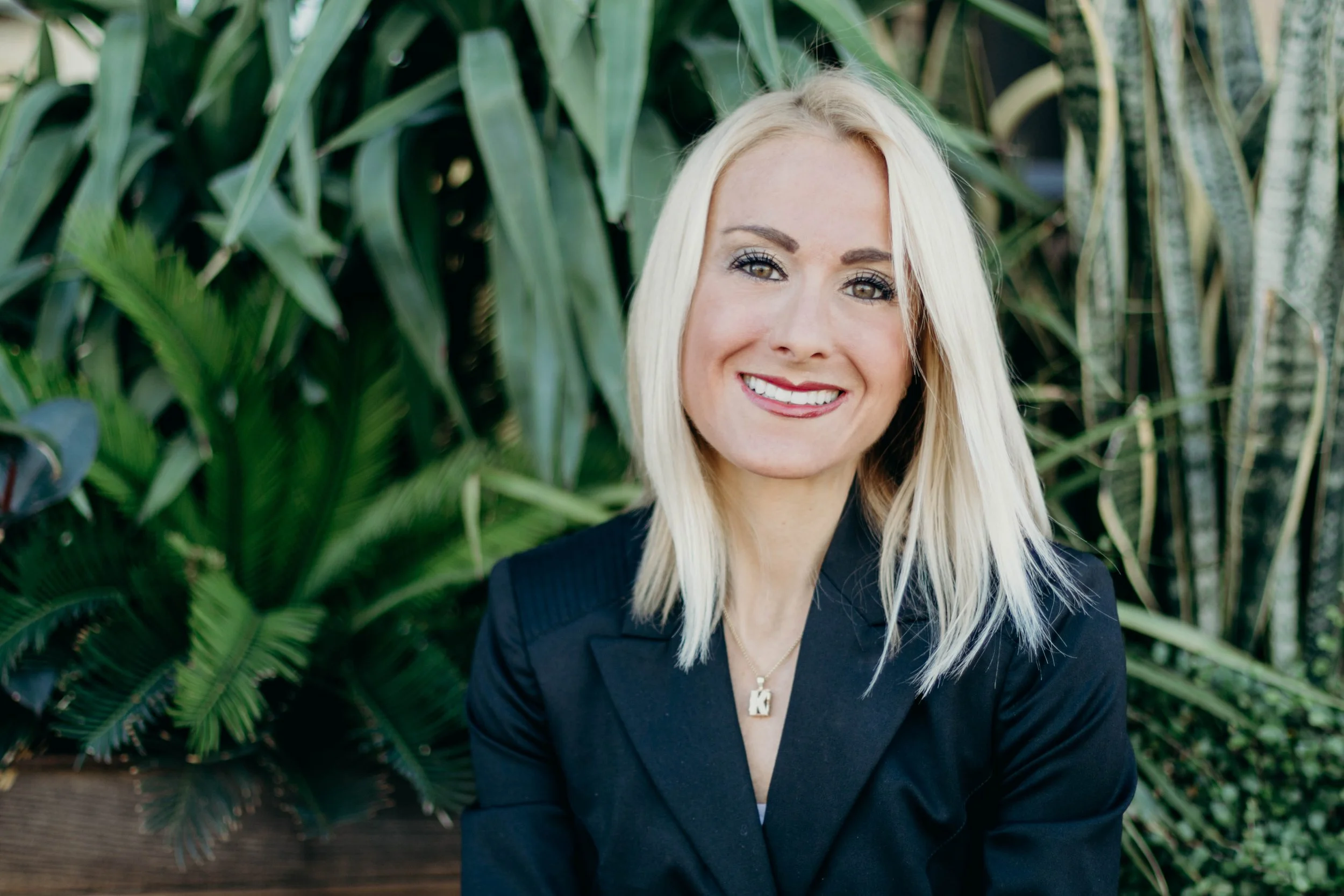 Headshot of public relations professional Katie Brashear in a black blazer and a necklace in front of lush green plants.