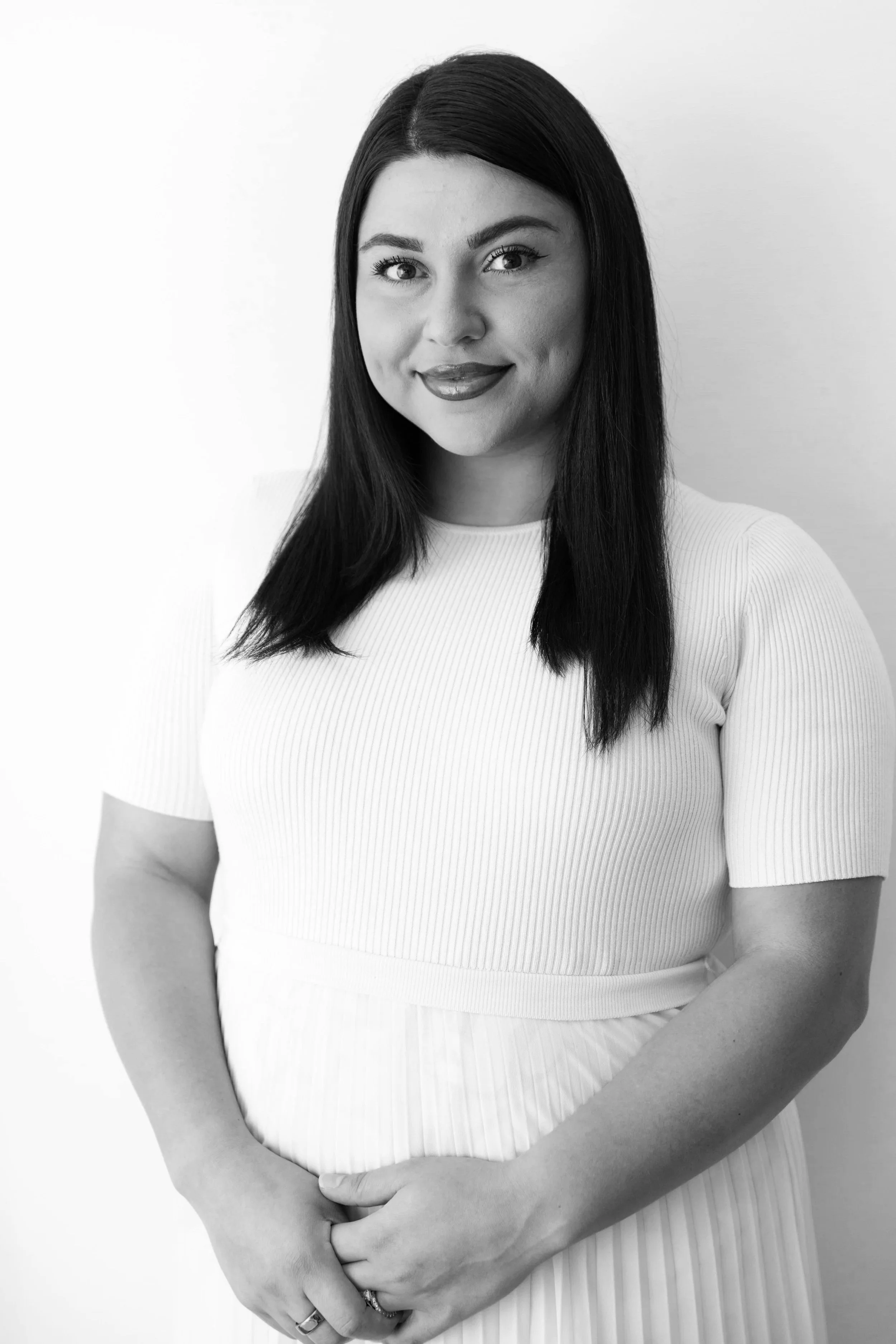 Black and white portrait of a woman with long dark hair, wearing a white ribbed dress, smiling gently, standing against a plain light background.