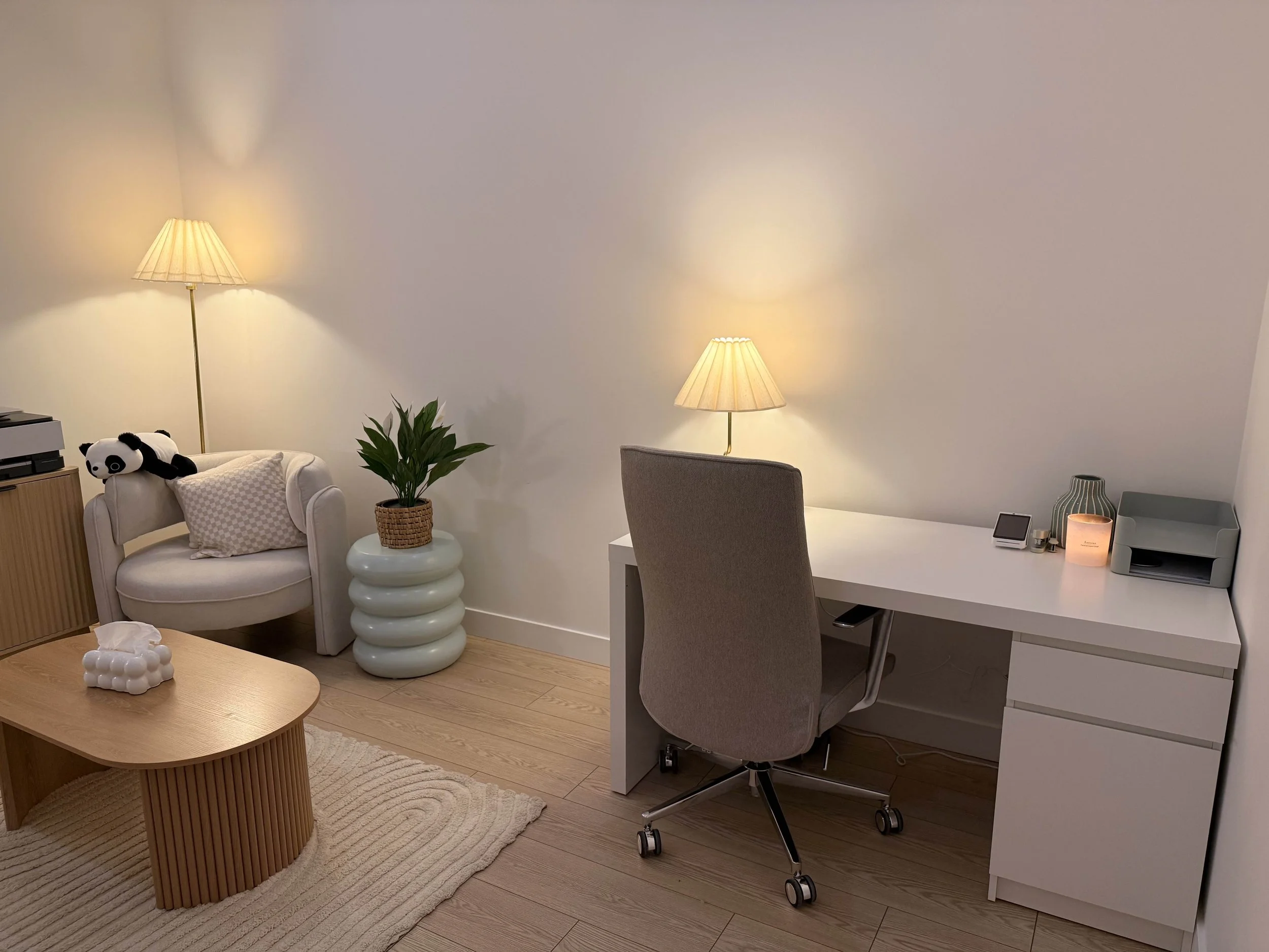 A cozy home office with a white desk, gray swivel chair, table lamp, decorative vase, and storage box, adjacent to a seating area with a plush armchair, a panda stuffed animal, and a potted plant.
