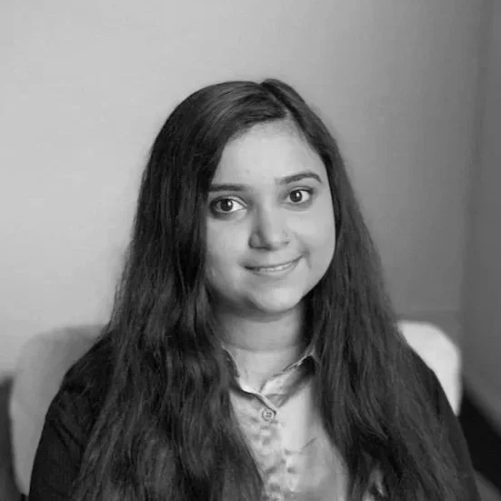 Black and white photo of a young woman with long wavy hair, smiling, wearing a collared shirt and blazer, sitting indoors.