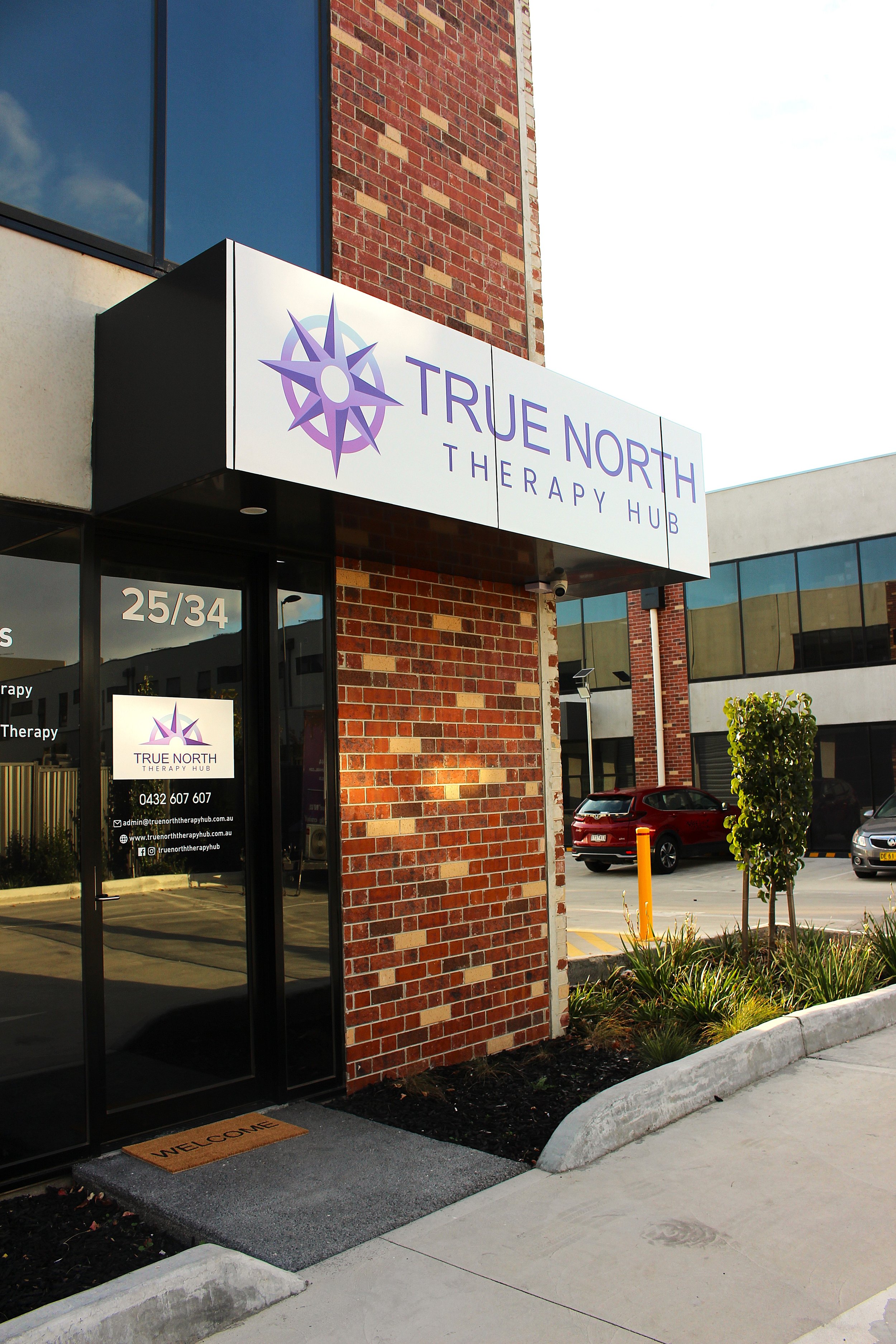 Exterior of a therapy center called True North Therapy Hub, with a sign featuring a purple compass logo. The entrance has a glass door with the center's contact information and social media handles. The building has a brick and glass facade, with a small, landscaped area and parked cars nearby.