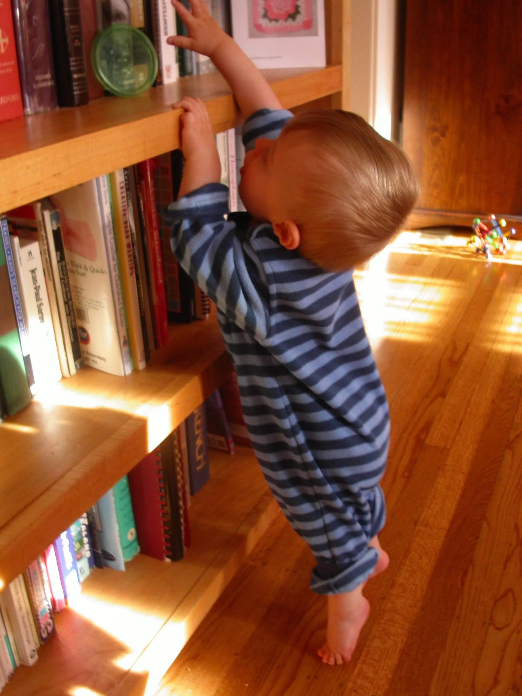 a toddler wearing a stripped onesie stands at a bookshelf reaching for a book.