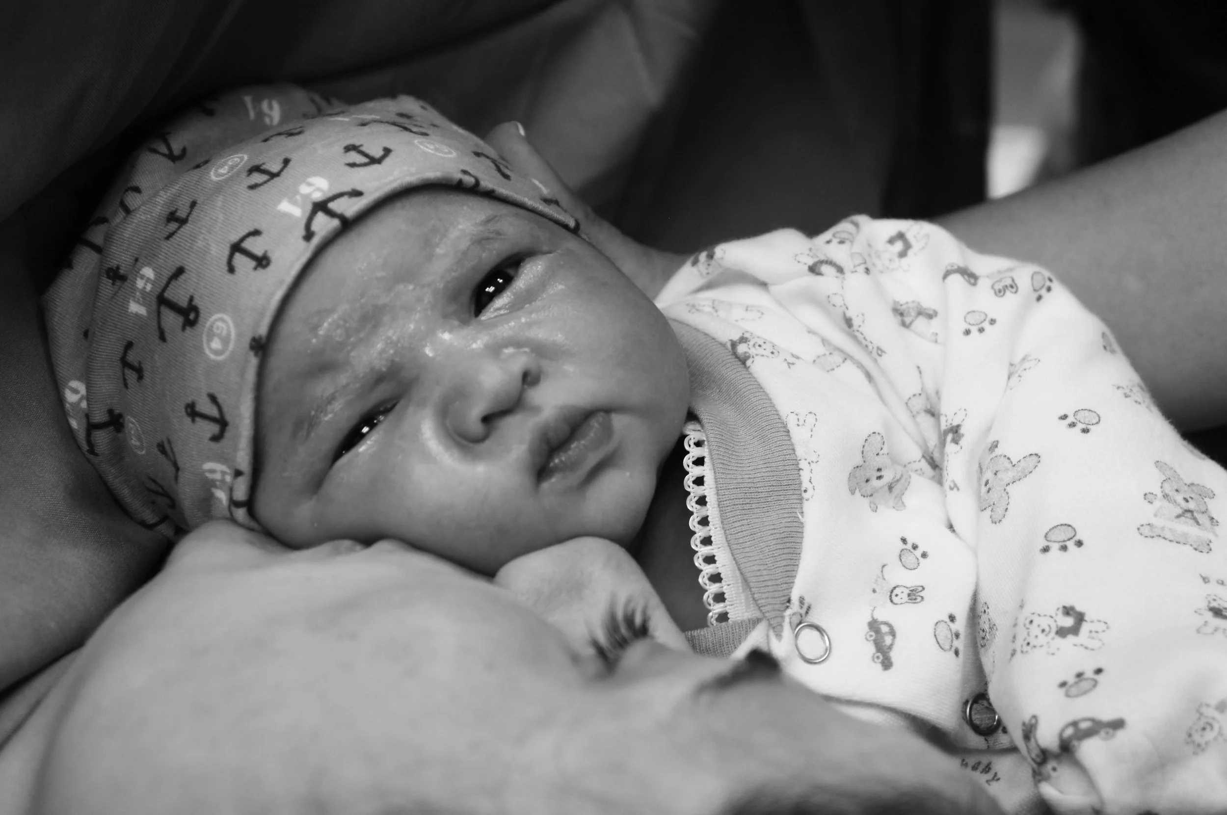A close-up, black and white photo of a freshly born baby receiving a kiss from their mother, wearing a hat with anchor patterns and a onesie with small bear designs.
