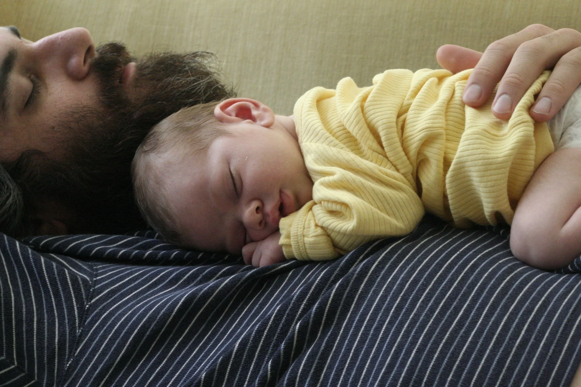 A new father with a beard and mustache and eyes closed lying on a bed while holding a sleeping baby dressed in a yellow outfit.