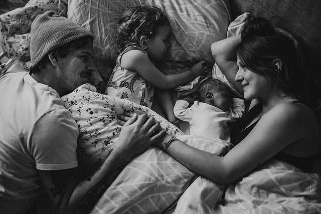 A black and white photo of a family lying in bed together, including a man and woman smiling at each other while holding hands across a sleeping young child and a baby, all smiling.