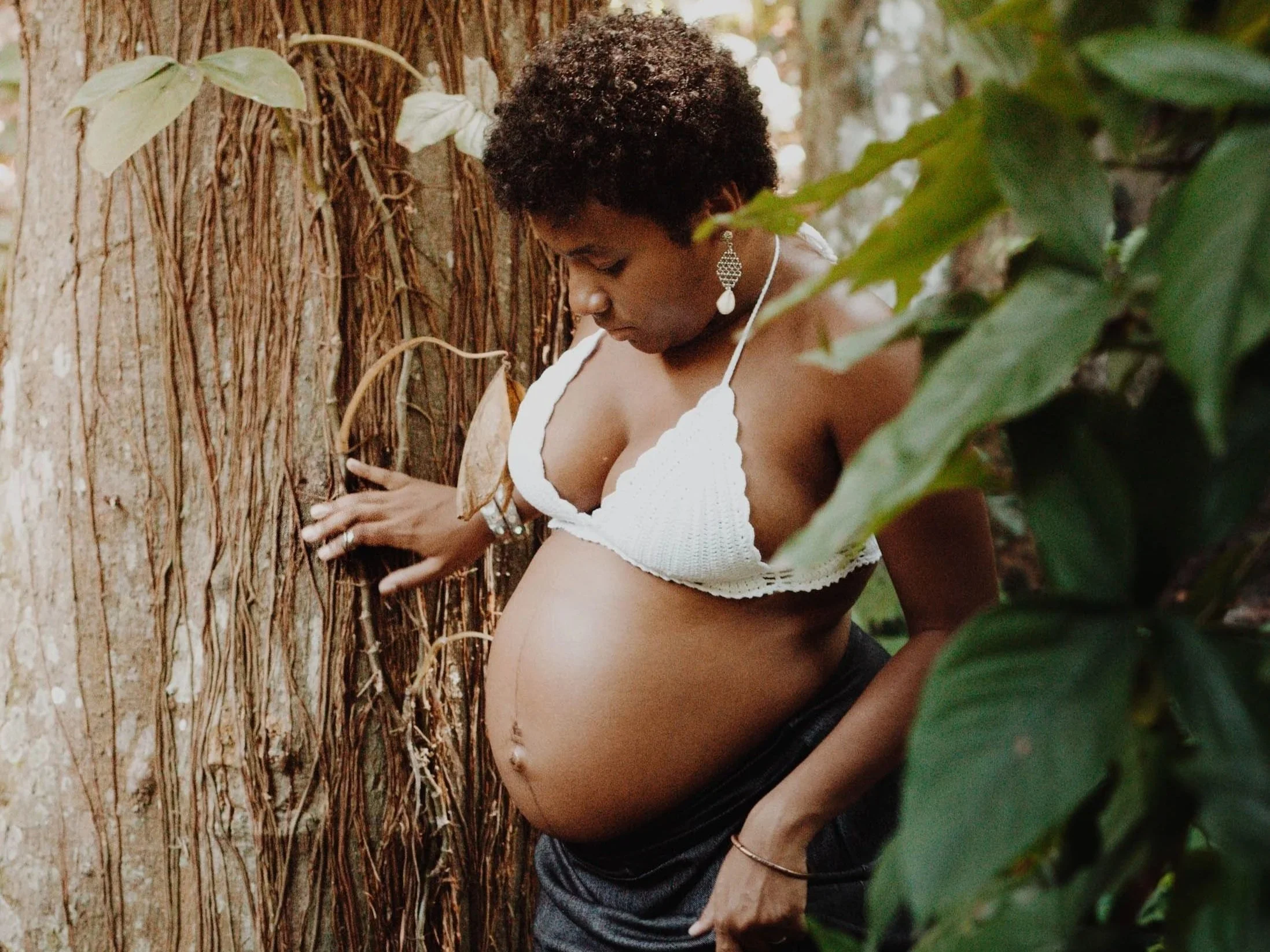 A woman with short curly hair, wearing a white crochet top and large earrings, is touching a large tree trunk with her right hand, surrounded by green leaves and foliage.