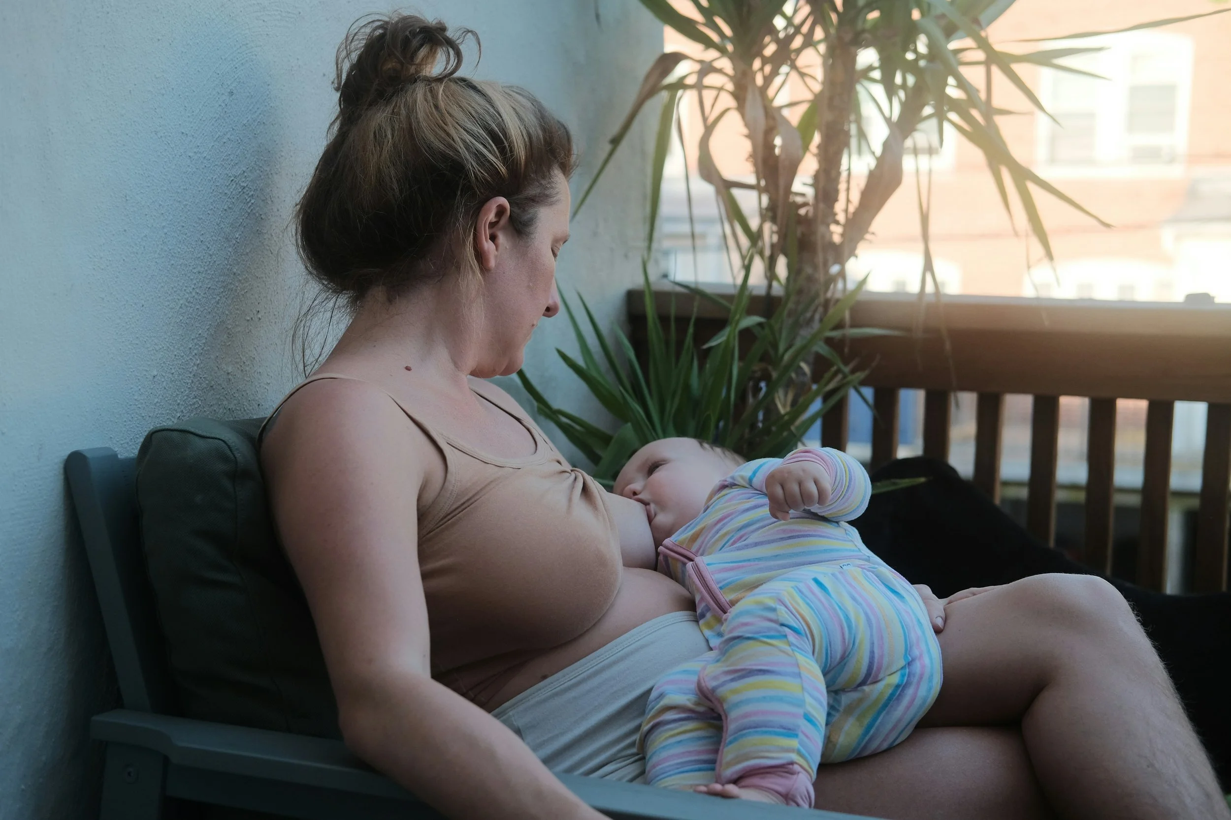 A woman gazes at her baby as she breastfeeds while sitting on a patio bench near a potted plant, with sunlight coming through a window in the background.