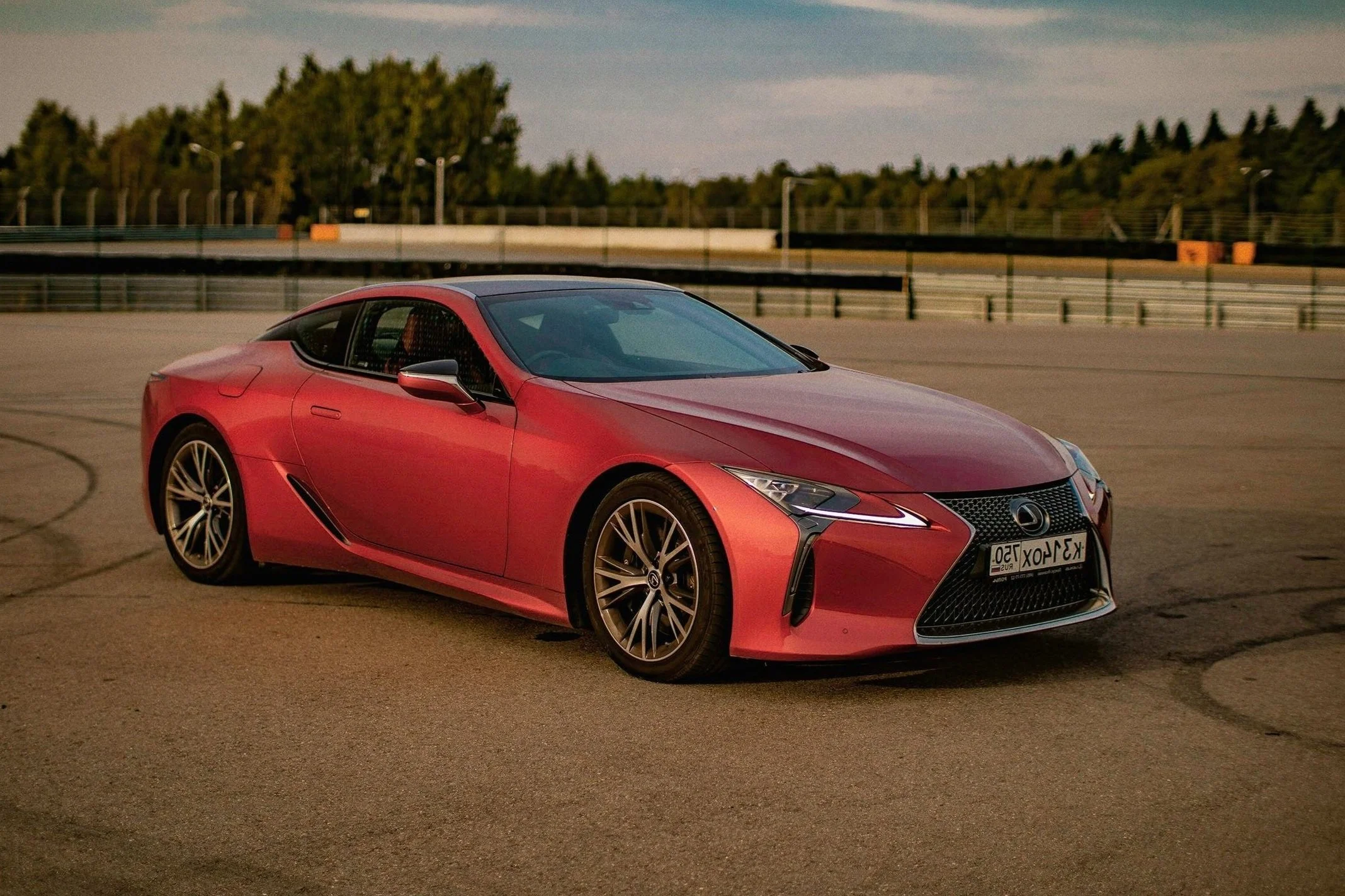 Red Lexus coupe parked on a racetrack with a blurred background of trees.
