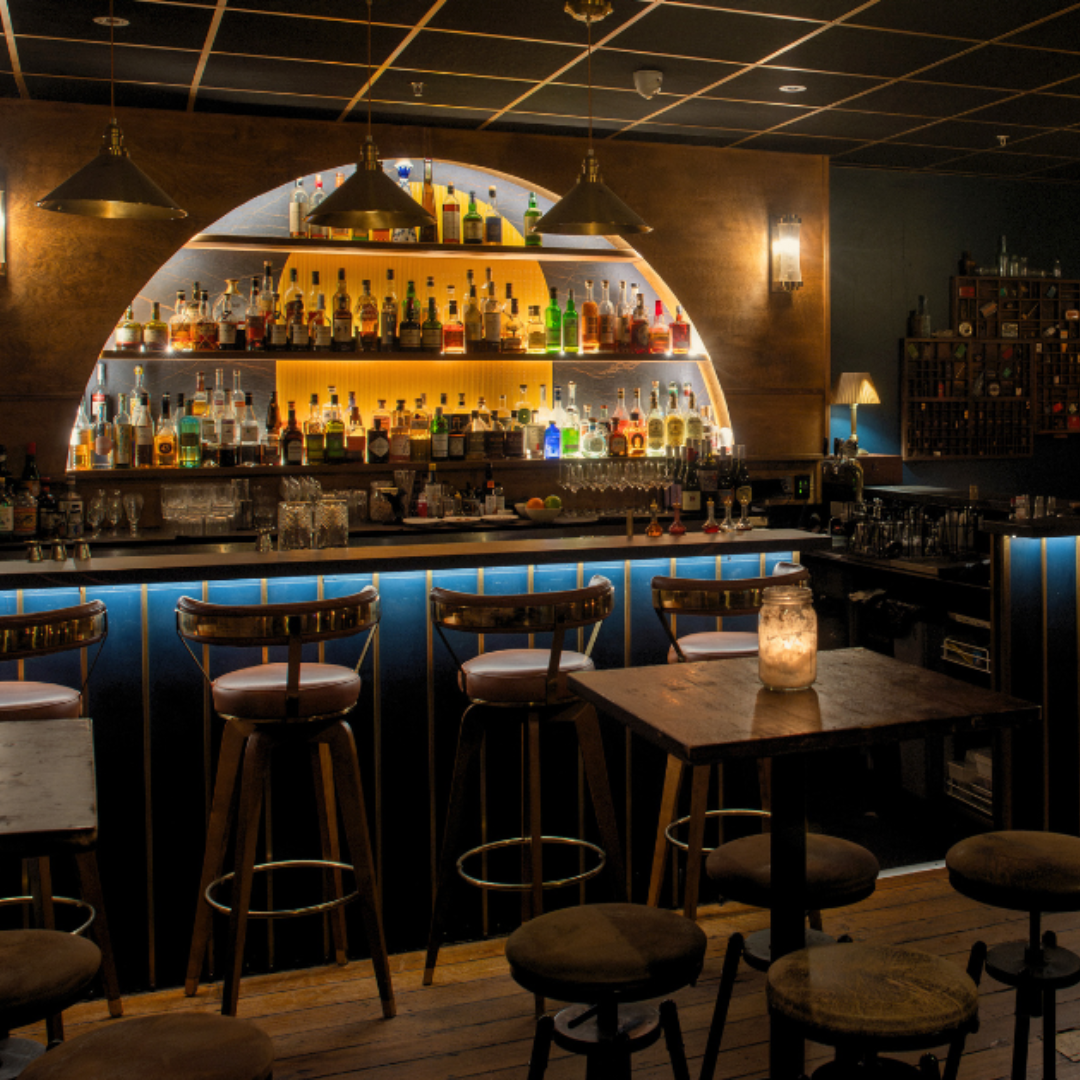 Dimly lit cocktail bar with jar candles on round wooden tables, leather seating, and framed art on the walls. Great date spot 