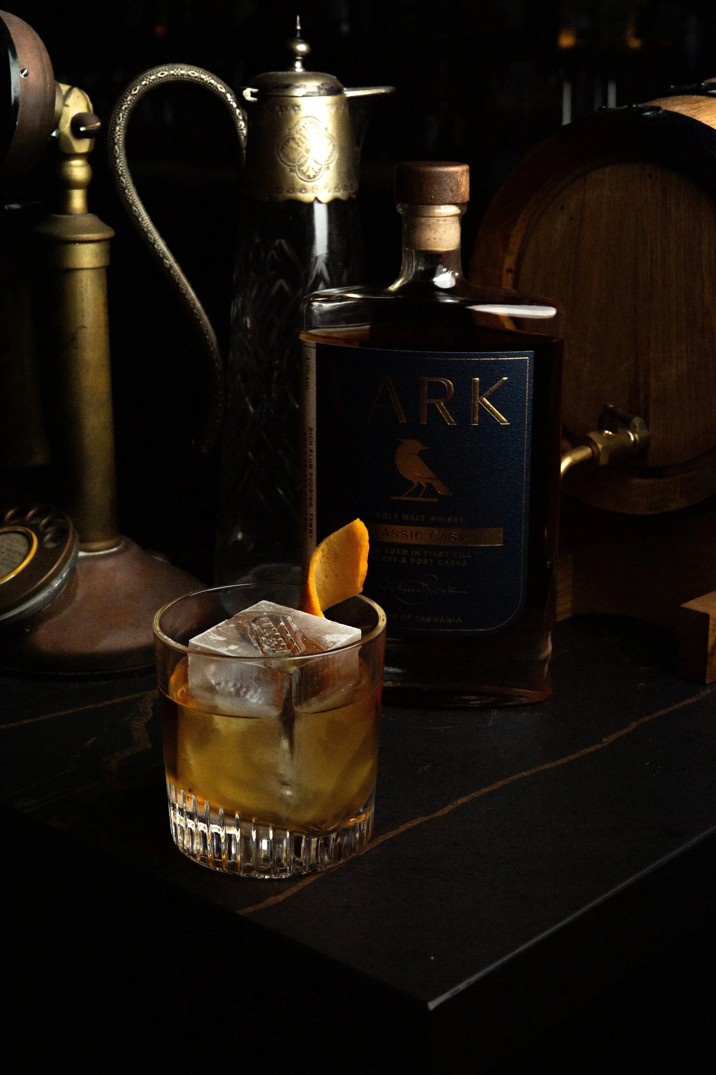 A glass of whiskey with ice and an orange peel garnish on a dark wooden table, with bottles and bar decor in the background.