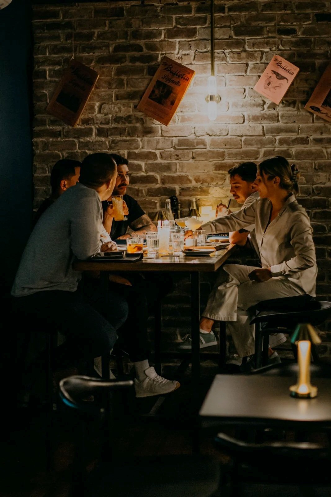 Cozy bar interior with a dimly lit atmosphere, featuring a semi-circular illuminated shelf lined with bottles and glasses. Five high stools are positioned at the bar counter, with additional tables and stools in the foreground. Brick walls and candles contribute to the warm ambiance.