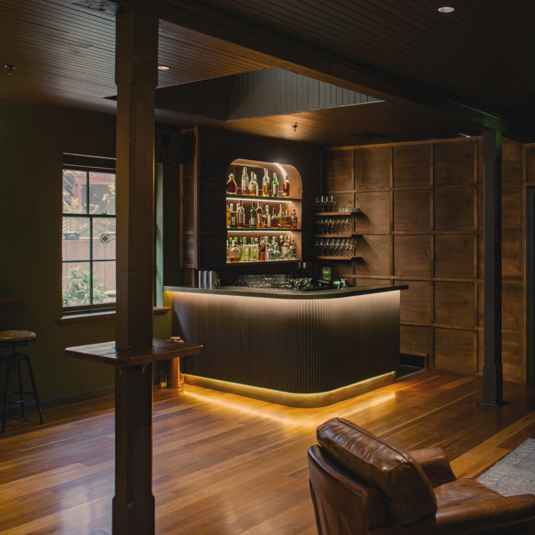 A dimly lit bar interior with a wooden bar counter, green walls, leather couches, and a rug on wooden flooring.
