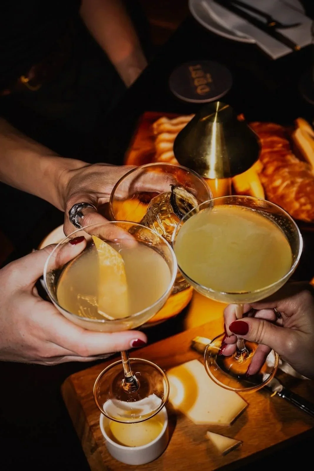 Three people toasting with cocktails in stemmed glasses over a tray with cheese and a sauce dish, with dim, warm lighting.