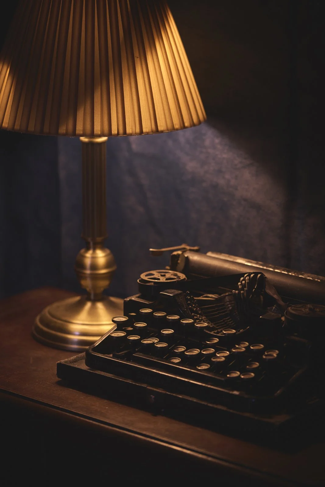Vintage typewriter on a wooden desk illuminated by a brass table lamp with a pleated shade.