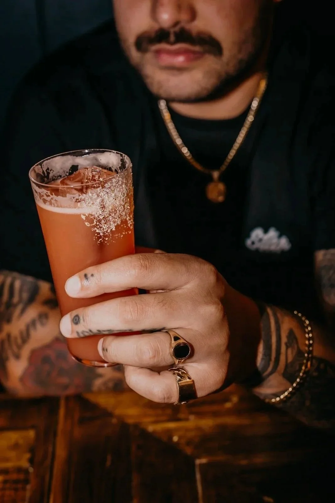 Hand holding a glass of red cocktail with smoke rising, against a background of vintage coasters on a wall.