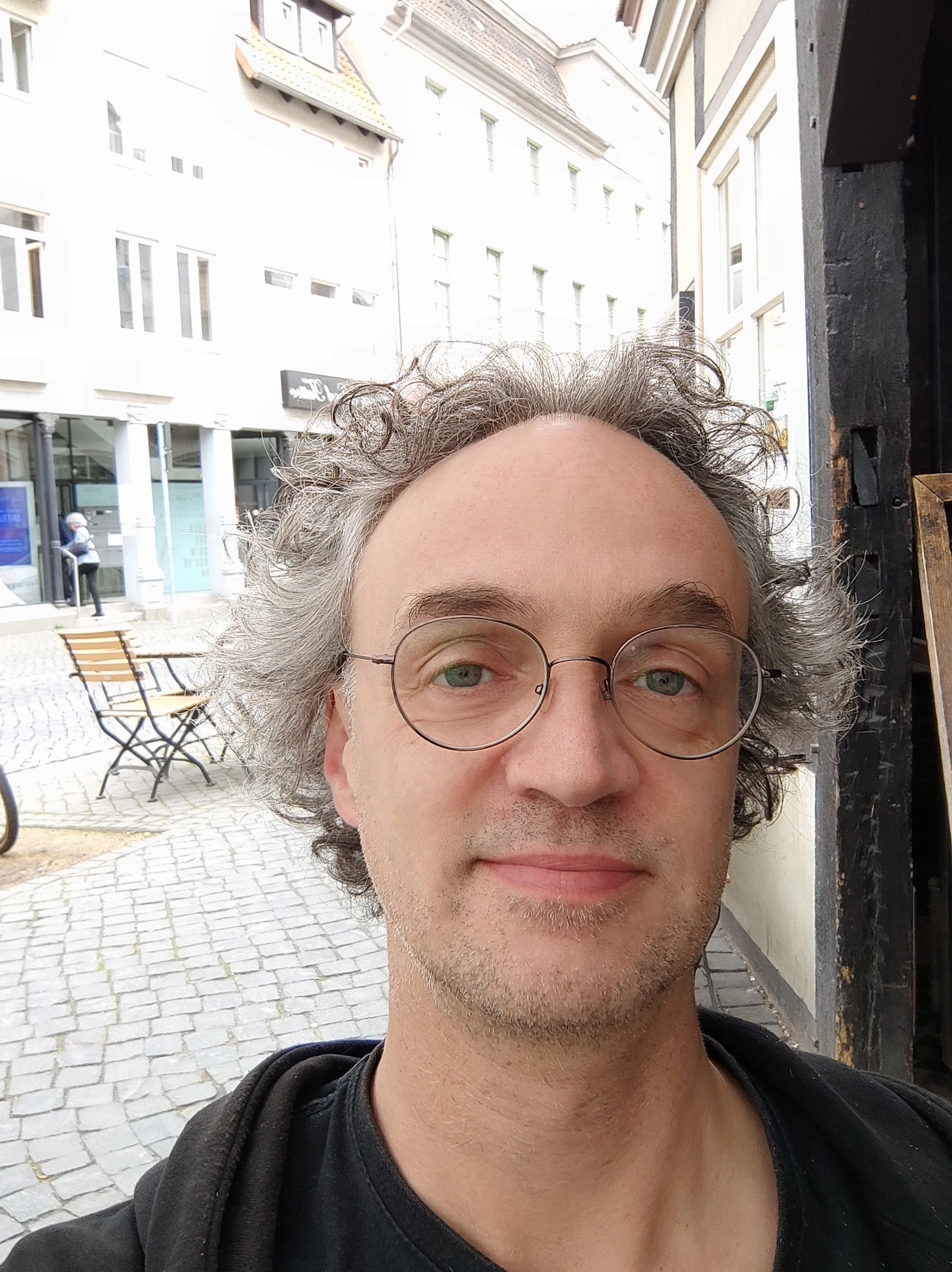 Ein Mann mit Brille und lockigem Haar macht ein Selfie auf einer Kopfsteinpflasterstraße in einer Stadt mit alten Gebäuden.
