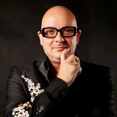 Uomo con testa calva, occhiali neri, giacca nera con dettagli decorativi dorati, che tocca il mento con la mano e indossa un anello dorato.