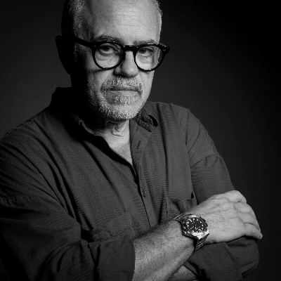 Uomo anziano con barba e occhiali, che indossa una camicia scura, con le braccia incrociate, in un'immagine in bianco e nero.