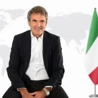 Uomo sorridente in abito nero con camicia bianca, sfondo con bandiera italiana.