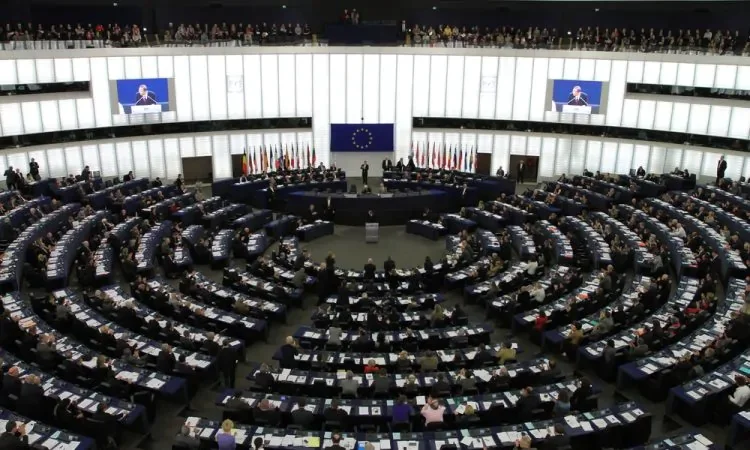 Sala plenaria del Parlamento Europeo con membri seduti a vari tavoli circolari, bandiere dei paesi membri e schermi con un oratore.