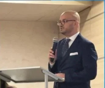 Uomo in abito elegante che parla al microfono durante un discorso o una presentazione. Sta dietro un podio in un ambiente interno.