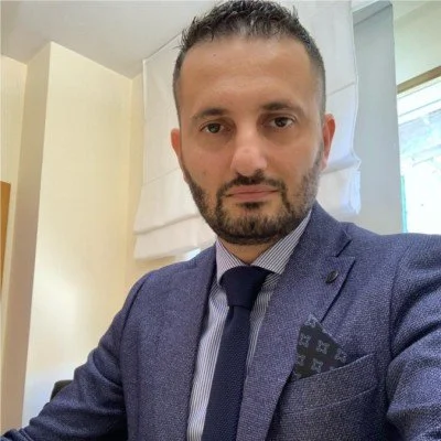 Uomo in abbigliamento formale con giacca blu, camicia a righe e cravatta, seduto in un ambiente interno con pareti chiare e una finestra.