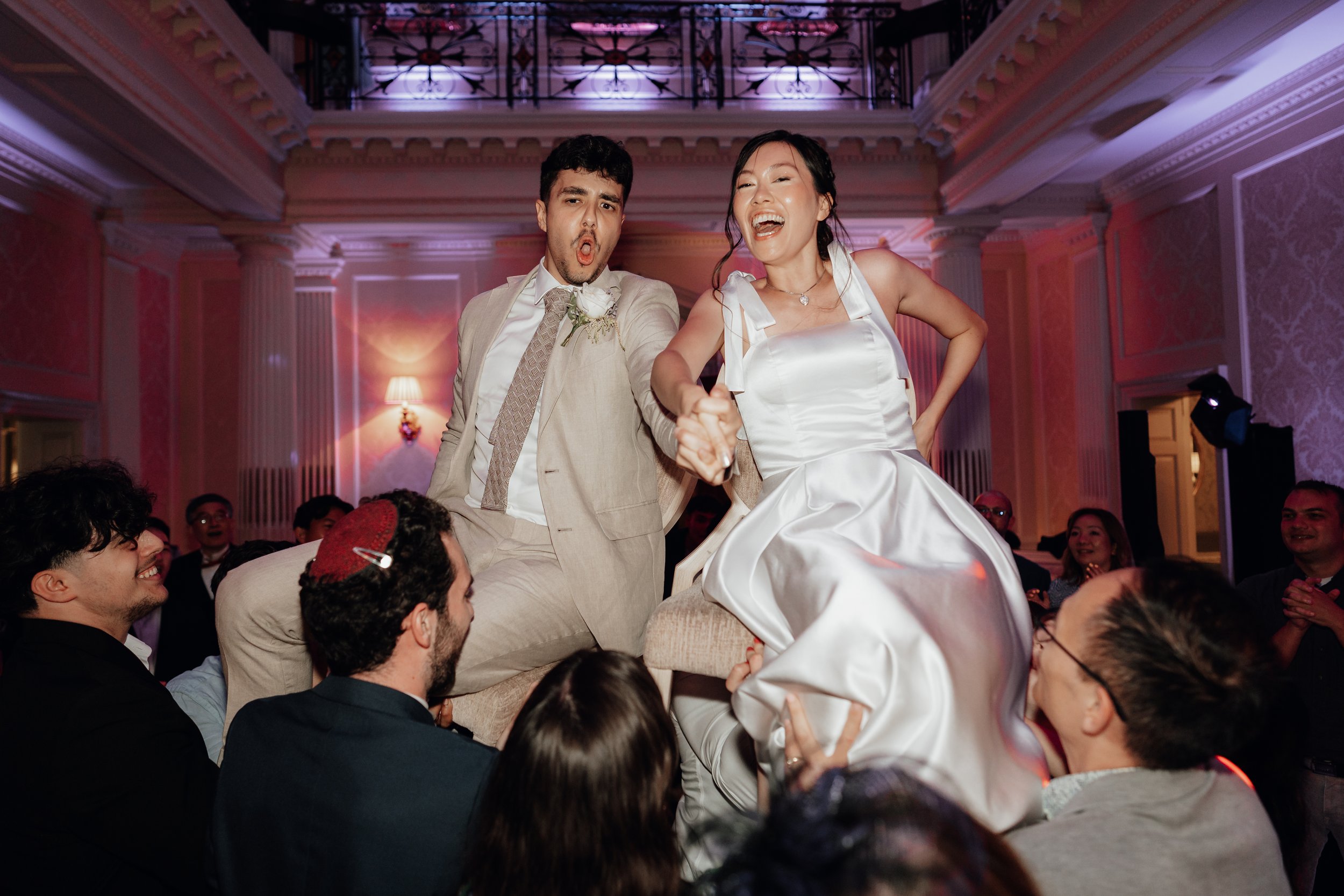 A bride and groom are lifted on chairs by guests during a wedding celebration. The bride is wearing a white dress and the groom a beige suit. Both are smiling and holding hands in a lively indoor setting.