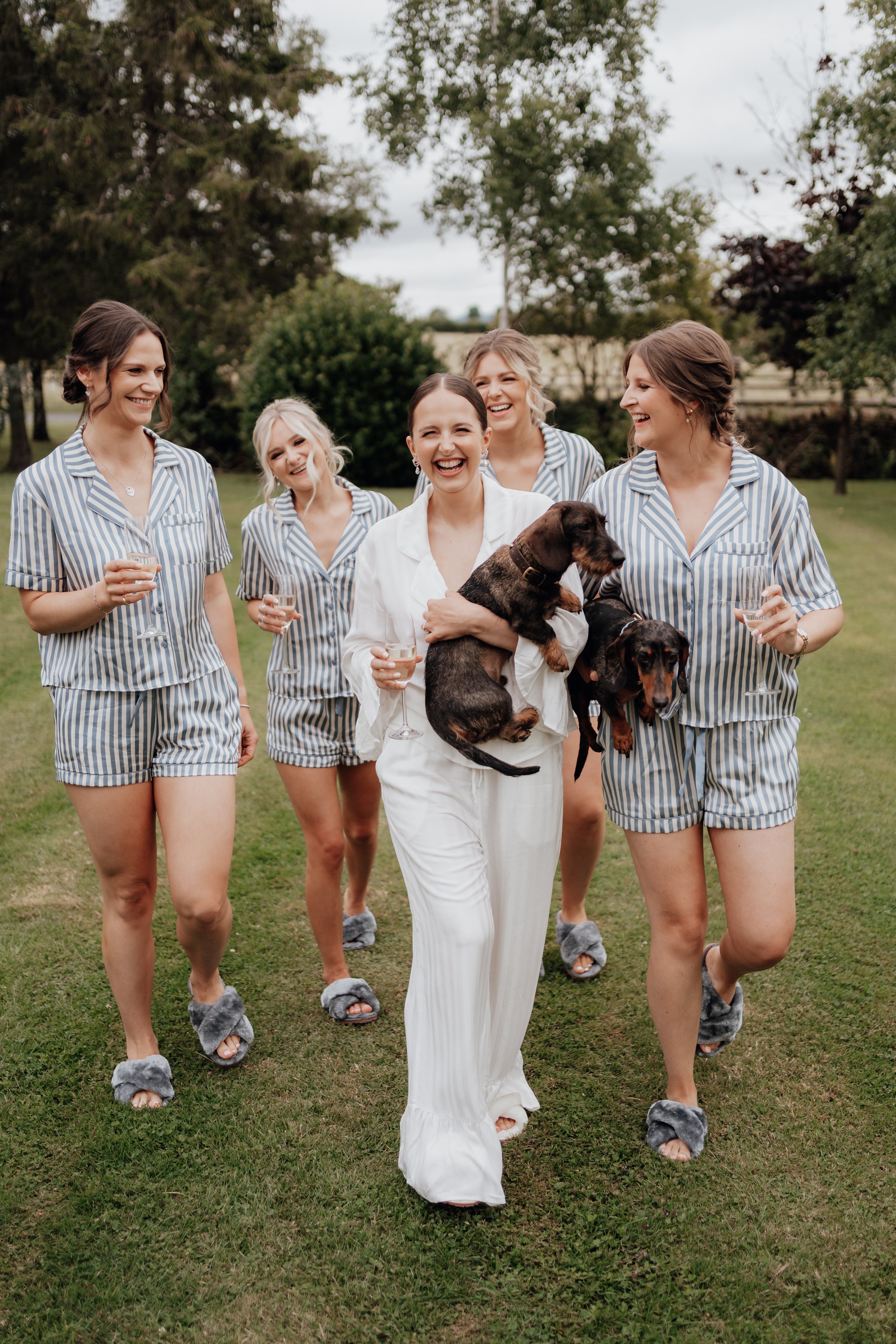 A group of women wearing matching striped pajamas, smiling and walking with two small dogs on green grass.