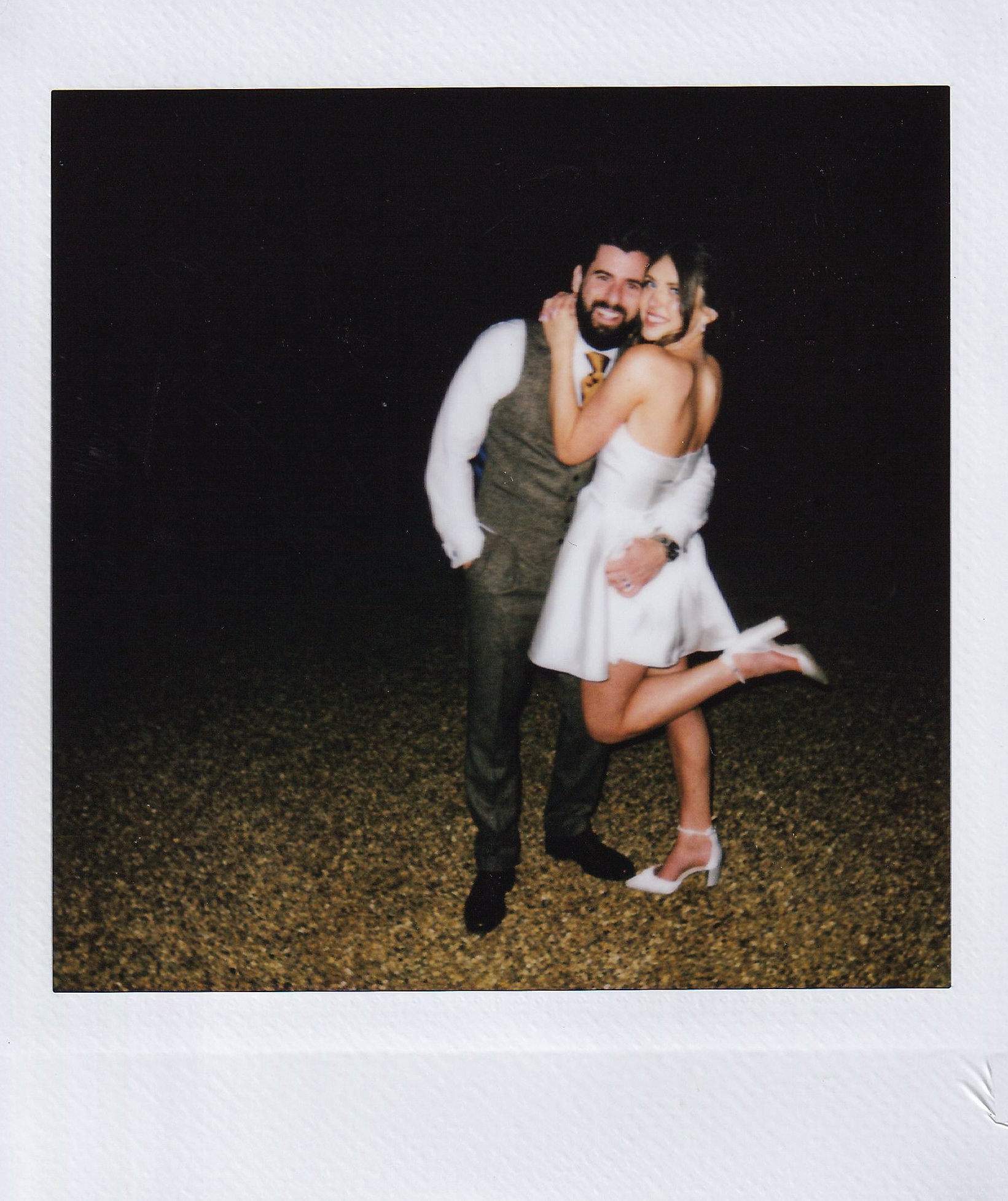 A couple poses together, dressed in formal attire. The man is wearing a suit with a vest, and the woman is wearing a short white dress with heels, lifting one leg playfully. They appear happy and are hugging each other.