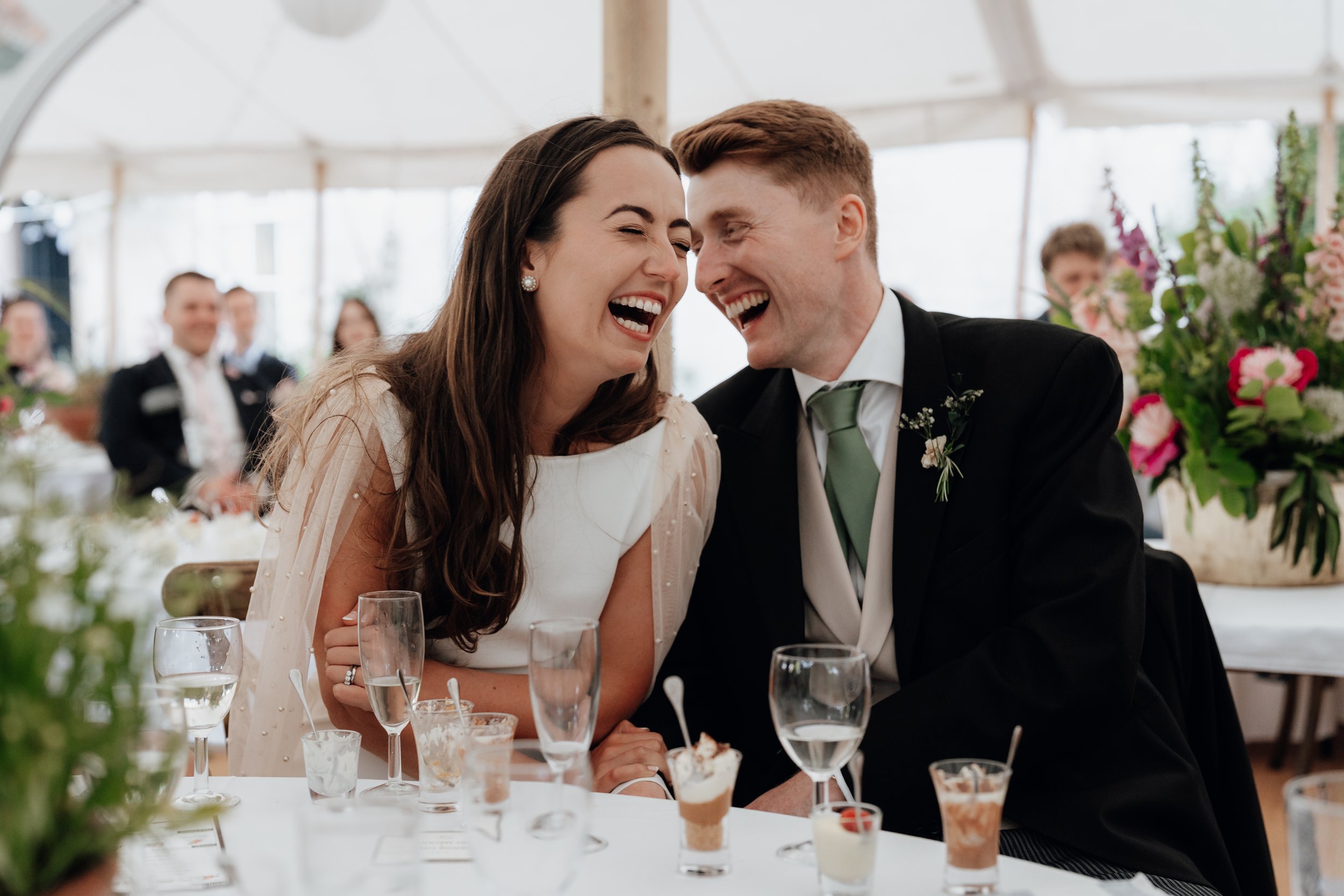 A bride and groom laughing at a wedding reception with elegantly decorated tables and guests in the background.