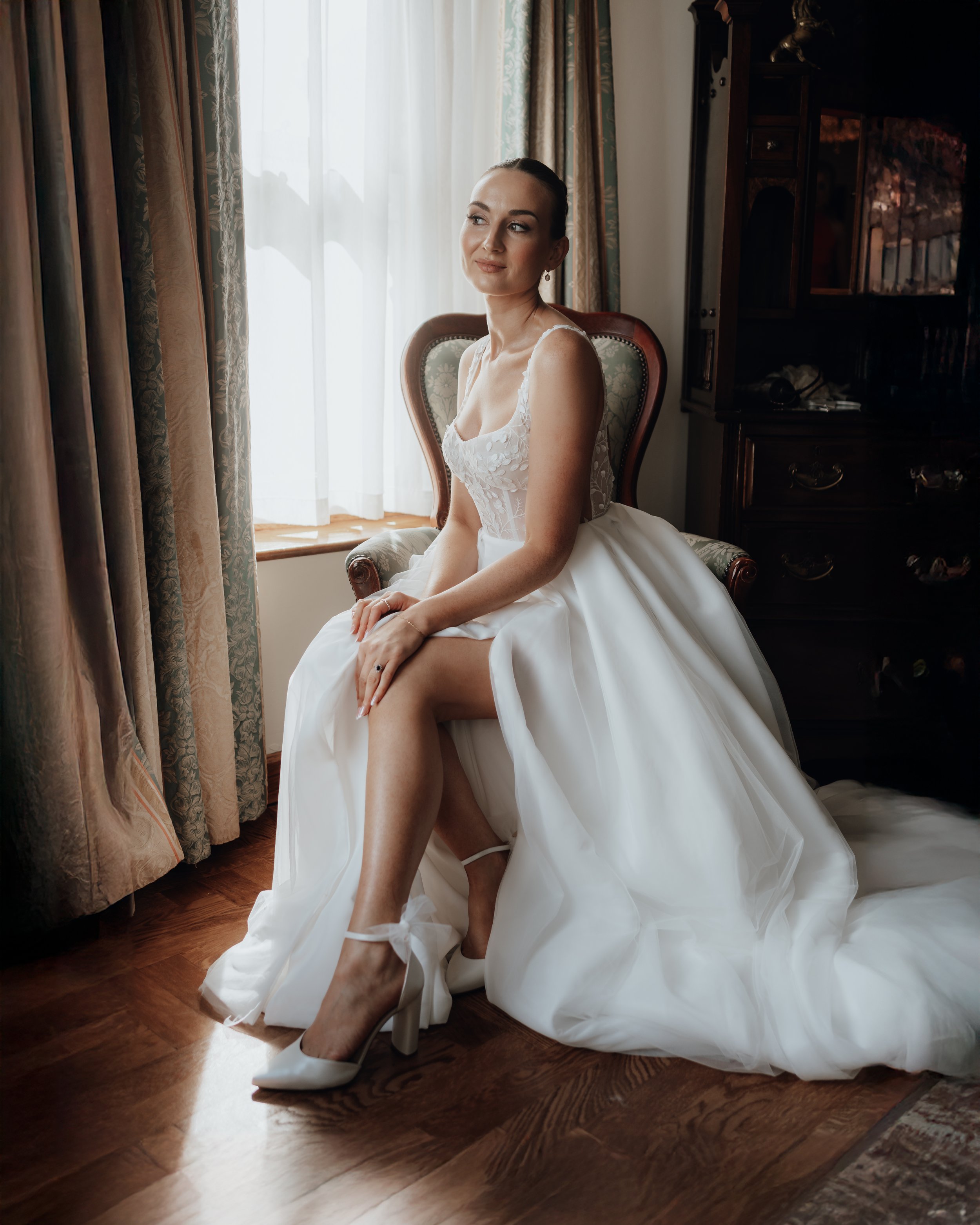 Bride in a white wedding dress sits on a vintage chair by a window, with light streaming through curtains, wooden floor visible.