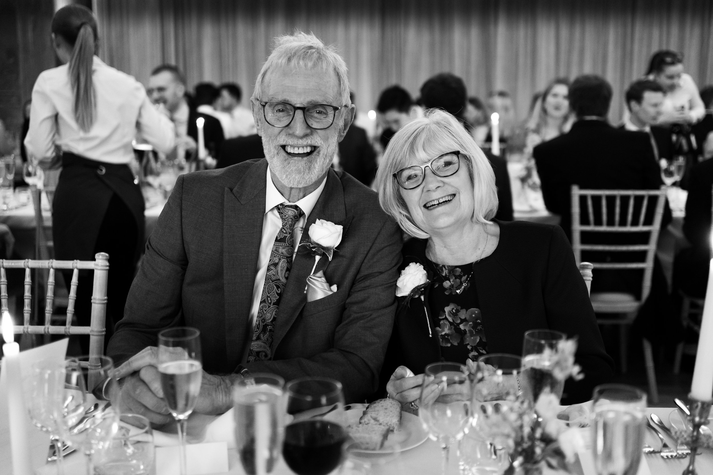 Elderly couple smiling at a formal event, seated at a table with flowers and wine glasses.