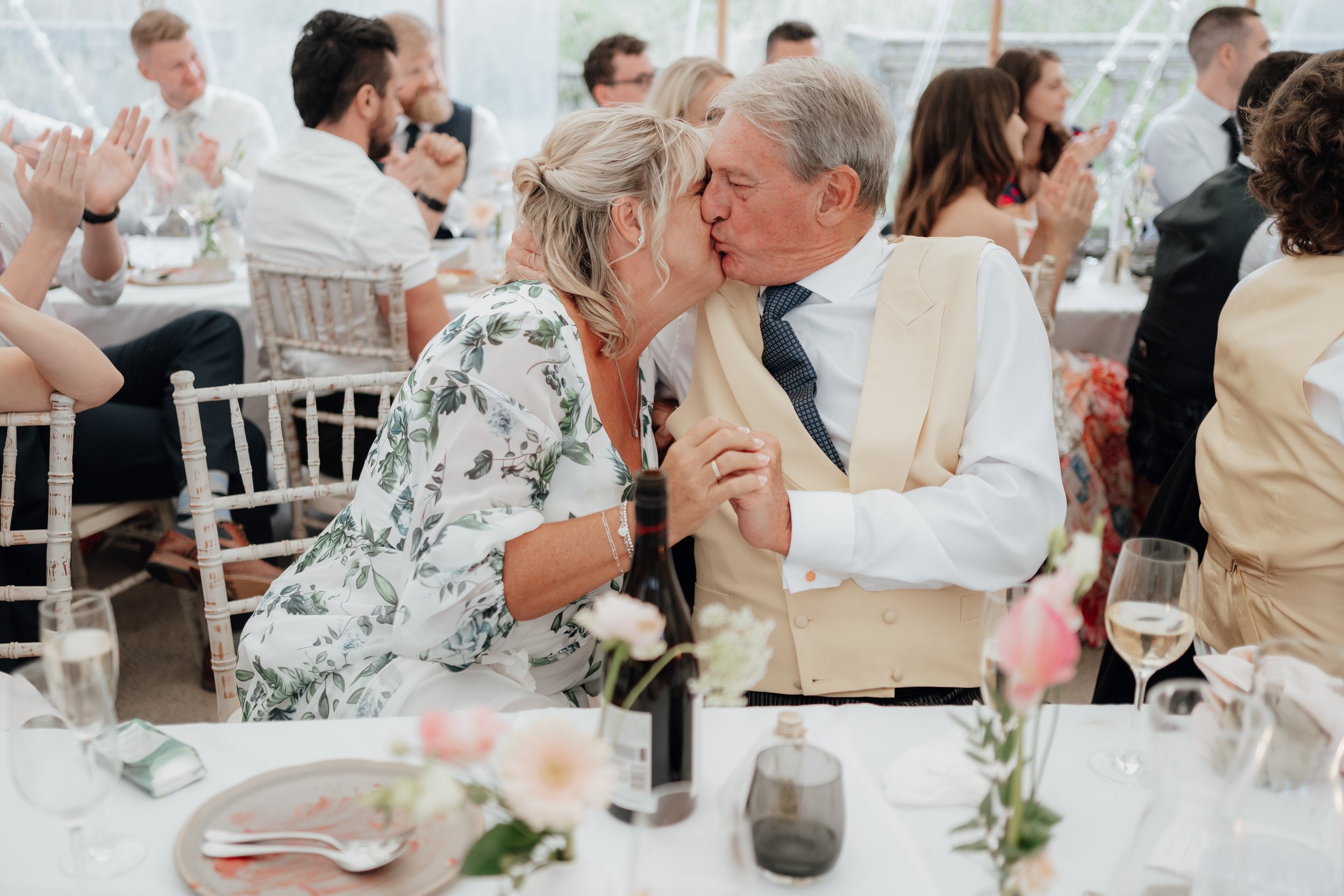 An elderly couple kissing at a formal event, surrounded by seated guests applauding. The table is elegantly set with flowers, wine, and glasses.
